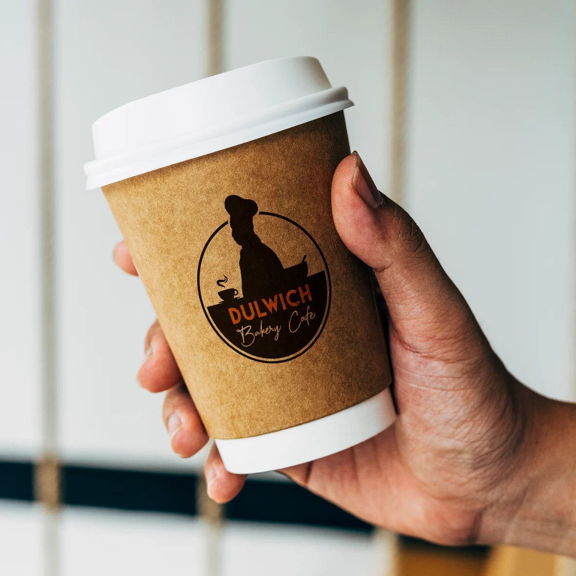 Hand holding a brown paper coffee cup with white lid and Dulwich Bakery logo