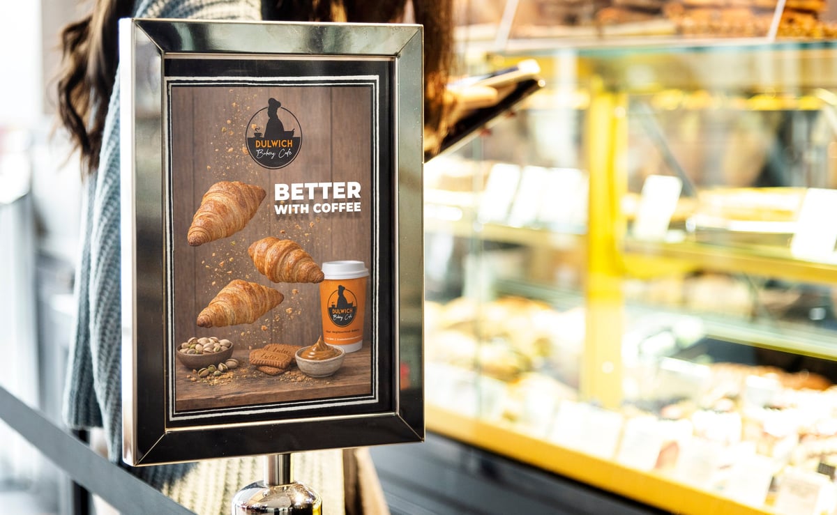 Framed bakery sign advertising fresh croissants and coffee, displayed outside a cafe with blurred yellow storefront in background